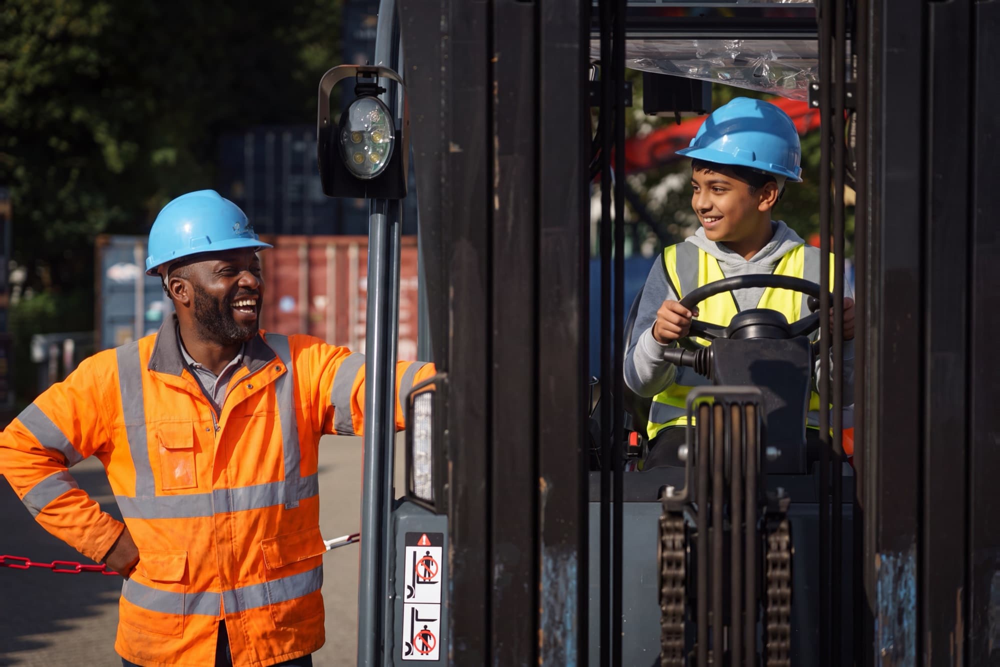 Een leerling achter het stuur van een heftruck, in gesprek met een lachende medewerker in oranje werkkleding.